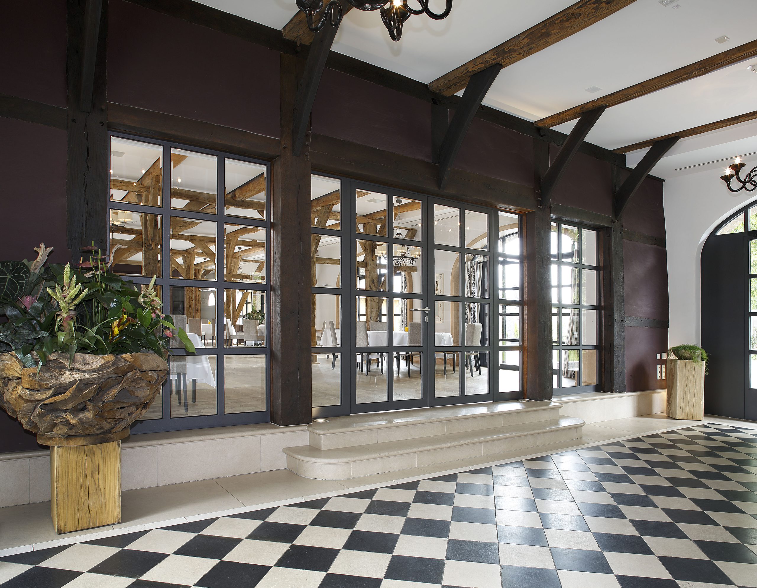Elegante Lobby mit schwarz-weiß-kariertem Boden, großen Fenstern, Holzbalken und Topfpflanzen auf Säulen.