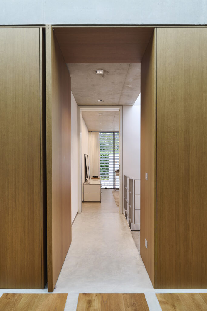 Blick durch eine Holztür in ein modernes, minimalistisches Schlafzimmer mit weißen Wänden und großen Fenstern.